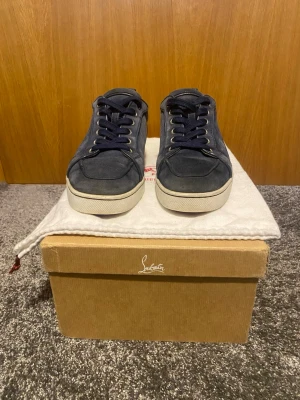 Marinblå sneakers från Louboutin - Snygga marinblå sneakers från Christian Louboutin i mocka med klassiska röda sulor. Skorna har vita kanter runt sulan, mörkblå skosnören och metallfärgade öljetter. Perfekta för dig som vill ha en exklusiv touch på din outfit. ÄKTA!