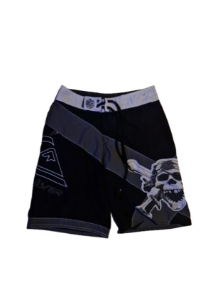 Svarta Quiksilver shorts med dödskalle - Snygga svarta shorts från Quiksilver med gråa detaljer och ett stort dödskalletryck på ena benet. Shortsen har snörning i midjan och är tillverkade i ett snabbtorkande syntetmaterial, perfekta för strand eller bad.