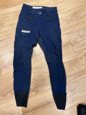 Marinblå ridbyxor från animo - Snygga marinblå ridbyxor från animo med svarta förstärkningar vid knäna och bensluten. Byxorna har två fickor fram med lock och detaljer i svart. Perfekta för ridning och träning.