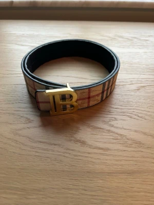 Burberry rutigt bälte med guldspänne - 105 cm, Stilrent bälte från Burberry med klassiskt rutig design i beige, rött och svart. Bältet har ett stort guldfärgat B-spänne framtil. Perfekt accessoar för att lyfta din outfit.