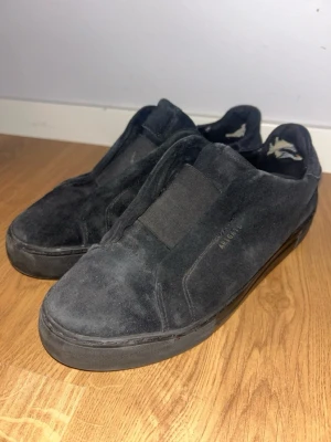 Svarta slip-on sneakers från Axel Arigato  - Stilrena svarta slip-on sneakers från Axel Arigato i mocka med elastiska band över vristen för enkel påtagning. Skorna har en låg profil och svart sula, perfekt för en clean och avslappnad look. Diskret logga på sidan och mjukt innerfoder.