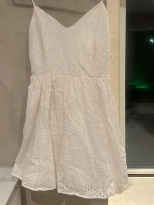 Vit kort klänning från H&M - Söt vit kort klänning från H&M med broderade hål och smala axelband. Klänningen har v-ringning och en lätt utställd kjol, perfekt för varma dagar. Tillverkad i bomull för en luftig och bekväm känsla.