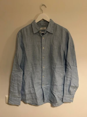 Ljusblå linneskjorta från Uniqlo - Säljer en stilren ljusblå skjorta från Uniqlo i svalt linne. Skjortan har klassisk krage, långa ärmar och vita knappar framtill. Perfekt för dig som gillar en clean och avslappnad look.