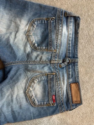 Blå jeans från ONLY med slitningar - Säljer ett par blå jeans från ONLY med snygga slitningar och detaljerade sömmar. Jeansen har fem fickor, dekorativa nitar och en klassisk knappgylf. Materialet är denim i bomull och passformen är rak med låg midja.