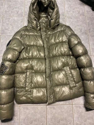 Olivgrön dunjacka från superdry  - Säljer en olivgrön dunjacka från superdry med glansig finish och stor huva. Jackan har två framfickor med lock och en superdry-logga på vänster ärm. Puffig modell som håller dig varm under kalla dagar. Perfekt för dig som vill ha både stil och funktion.