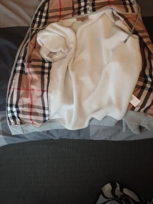 Burberry hoodie med klassiskt rutmönster - Snygg hoodie från Burberry i storlek M. Utsidan har det ikoniska beige, svarta och röda rutmönstret och insidan är mjukt vitt fleece-material. Hoodien har huva med dragsko och ribbade muddar. Perfekt för dig som vill ha en exklusiv och trendig look.
