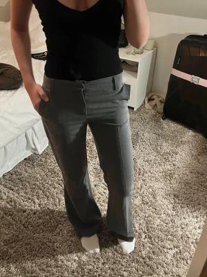 Grå kostymbyxor med raka ben - Snygga grå kostymbyxor med raka ben och pressveck. Byxorna har normalhög midja, hällor för skärp och sidfickor. Perfekta för en clean och stilren look. Materialet ser ut att vara en mjuk och lätt blandning, troligen polyester eller viskos.