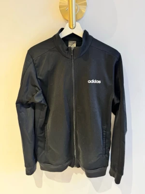 Svart ziptröja från Adidas - Svart ziptröja från Adidas med vit logga på bröstet. Tröjan har hög krage, långa ärmar och två sidofickor. Tillverkad i ett mjukt och stretchigt material som passar perfekt till träning eller chill.