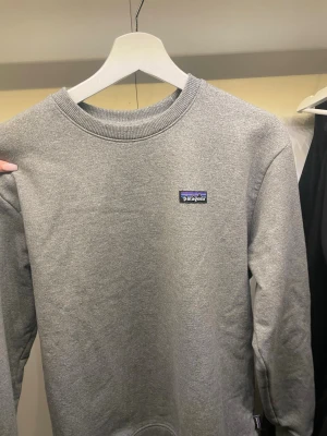 Grå sweatshirt från Patagonia - Klassisk grå sweatshirt från Patagonia med rund halsringning och diskret logga på bröstet. Tröjan har lång ärm och är tillverkad i mjuk bomullsmix som passar perfekt till vardagslooken. Enkel och stilren design som funkar till det mesta.