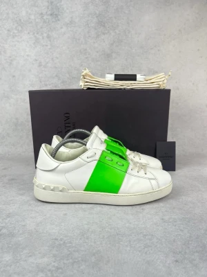 Valentino vita sneakers med grön detalj - Snygga vita sneakers från Valentino med en fet neongrön panel över snörningen och coola nitar på hälen. Skorna är låga och har en klassisk siluett i läder som ger en clean men ändå unik vibe. Perfekta för dig som vill sticka ut med stil.