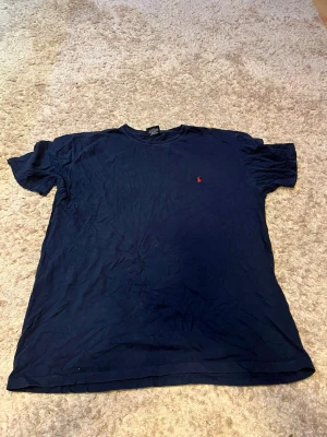 Mörkblå t-shirt från Polo Ralph Lauren - Klassisk mörkblå t-shirt från Polo Ralph Lauren med den ikoniska röda logon broderad på bröstet. T-shirten har rund halsringning och korta ärmar. Tillverkad i 100% bomull för en skön och avslappnad känsla.