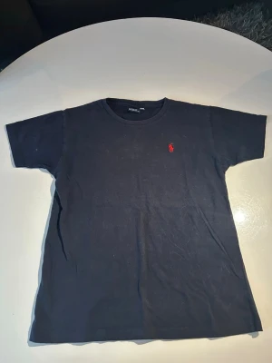 Mörkblå t-shirt från Polo Ralph Lauren - Klassisk mörkblå t-shirt från Polo Ralph Lauren med röd broderad logga på bröstet. T-shirten har rund halsringning och korta ärmar. Perfekt basic-plagg med stilren design och skön passform.