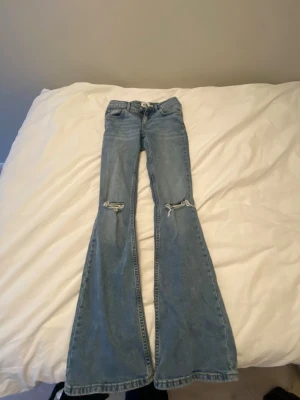 Blå utsvängda jeans från 157 - Säljer ett par ljusblå jeans från 157 med utsvängda ben och slitningar vid knäna. Klassisk femficksmodell med hög midja och bälteshällor. Jeansen är gjorda i denim och har en cool vintage vibe.