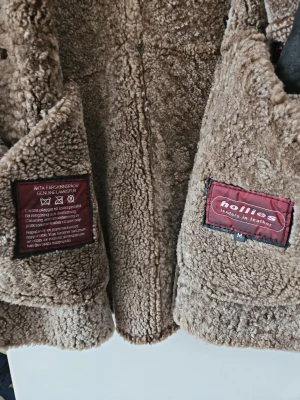 Hollies shearlingjacka i äkta lammskinn – strl M (mycket fint skick) - Säljer en exklusiv shearlingjacka från Hollies, tillverkad i 100 % äkta lammskinn med äkta fårskinnspäls på insidan. En varm, tidlös kvalitetsjacka i premiumsegmentet. Jackan är i mycket fint skick och i praktiken oanvänd, med ett mindre kosmetiskt fel (se nedan). Detaljer: Typ: Shearling / lammskinnsjacka Anmärkning: Ett litet snitt intill en knapp, mycket diskret och knappt synligt. Påverkar inte funktion eller hållbarhet.