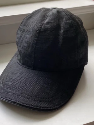 Svart keps från Fendi med logga - Snygg svart keps från Fendi med diskret FF-logga i tyget och justerbart spänne bak i läder. Klassisk böjd skärm och stilren design som passar till det mesta. Materialet känns exklusivt och kepsen har märkets taggar kvar.