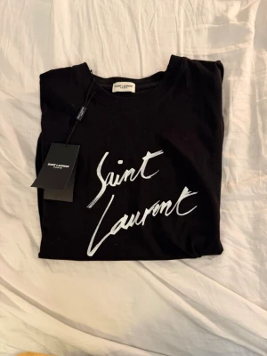 Saint Laurent t-shirt - Säljer en svart t-shirt från Saint Laurent med vit logga i handskriven stil på bröstet. Klassisk rund halsringning och korta ärmar. Tillverkad i mjuk bomull för en skön och avslappnad look.