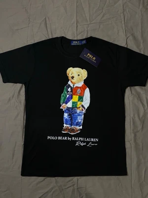 Svart Polo Bear t-shirt Ralph Lauren - Svart t-shirt från Ralph Lauren med ikoniskt Polo Bear-tryck framtill. Björnen har färgglada kläder och texten 'POLO BEAR by RALPH LAUREN' under motivet. Klassisk rund hals och korta ärmar. Perfekt för dig som gillar streetstyle och snygga prints.
