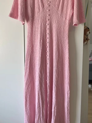 Ljusrosa långklänning med knappar - Säljer en ljusrosa långklänning med korta ärmar och knappar hela vägen framtill. Klänningen har en mjuk, flowy passform och är perfekt för varma dagar. Ingen synlig logga eller märke.