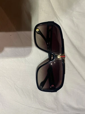 Svarta Carrera solglasögon  - Snygga svarta solglasögon från Carrera med bred båge och gulddetaljer vid näsryggen och på sidorna. Glasen är mörka och rektangulära, vilket ger en cool och modern look. Perfekta för att lyfta din stil under soliga dagar. Säljer för att de inte kommer till användning. Nypris är ca 2095kr, hör av er vid frågor samt om ni vill ha fler specifika bilder! 