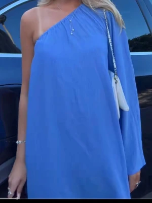 Blå oneshoulder klänning - Snygg blå oneshoulder klänning med lång ärm på ena sidan och bar axel på andra. Klänningen har en lös passform och är perfekt för sommarens alla tillfällen. Enkel och stilren design utan mönster eller extra detaljer. Finns små mindre rosa fläckar. Köpt på Vinted inget hon informerat om