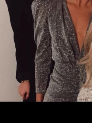 Nyskick second Female Mq Glittrig silver omlottklänning Stl s - Säljer en glittrig silverfärgad omlottklänning med v-ringning och långa ärmar. Klänningen har en figurnära passform och slutar ovanför knäna. Dold Knapp i v-ringning man kan knäppa eller uppknäppt beroende på urringning . Använd en gång