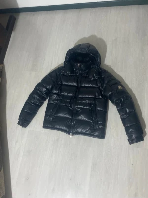 Svart pufferjacka från Moncler - Snygg svart pufferjacka från Moncler med glansig finish och klassisk Moncler-logga på ärmen. Jackan har huva, dragkedja framtill och två sidofickor. Fylld med dun för extra värme och har en quiltad design som ger en modern look.