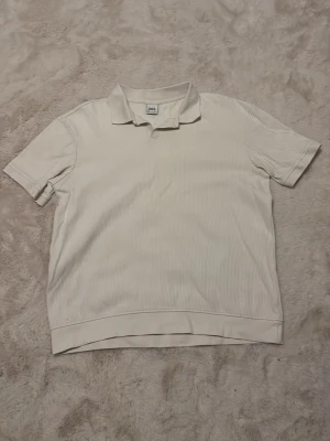 Vit ribbad polotopp från Zara - Säljer en vit ribbad polotopp från Zara i storlek M. Toppen har korta ärmar, klassisk krage och är tillverkad i ett mjukt material som känns skönt mot huden. Enkel och stilren design som passar till många olika outfits.