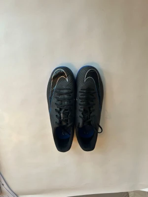 Nike Mercurial svarta fotbollsskor - Säljer ett par svarta Nike Mercurial fotbollsskor med vita och blå detaljer. Skorna har en låg profil, snörning och en sula som passar för spel på inomhusunderlag. Tillverkade i syntetmaterial med klassisk Nike Swoosh på ovansidan och Mercurial-tryck på hälen.