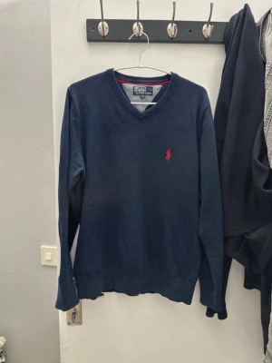 Mörkblå stickad tröja Polo Ralph Lauren - Mörkblå stickad tröja från Polo Ralph Lauren med V-ringning och röd broderad logga på bröstet. Tröjan har ribbade muddar vid ärmslut och nederkant. Har L som storlek men passar även M.