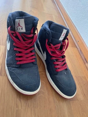 Air Jordan 1 mid denim blue 48.5 - Snygga Air Jordan 1 High sneakers i mörkblå canvas med vita swoosh-detaljer och röd Air Jordan-logga på sidan. Skorna har röda skosnören och vit sula med röd undersida. Perfekta för dig som gillar klassisk streetwear-stil.