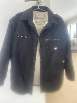 Svart jacka från Zadig & Voltaire - Stilren svart jacka från Zadig & Voltaire med vitt varmt teddyfoder på insidan. Jackan har klassisk krage, tryckknappar framtill, bröstficka med dragkedja och dekorativ logotyp på bröstet. Perfekt för lager-på-lager och en cool streetstil. Rymlig i storleken