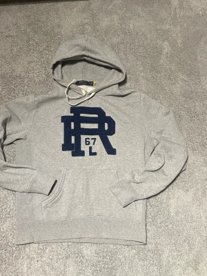 Grå hoodie från Polo Ralph Lauren - Snygg grå hoodie från Polo Ralph Lauren med stor mörkblå PR logga på bröstet. Klassisk känguruficka framtill, dragsko i huvan och ribbade muddar. Perfekt för en avslappnad och sportig stil.