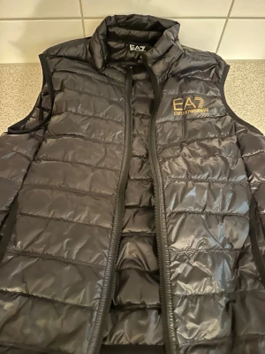 Svart dunväst från Emporio Armani - Snygg svart dunväst från Emporio Armani EA7 med guldfärgad logga på bröstet. Västen har hög krage, dragkedja framtill och quiltad design. Perfekt för lager på lager och ger en sportig, trendig look.
