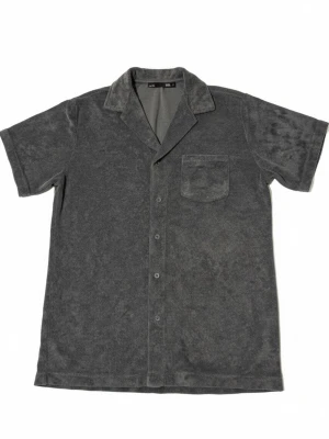 Grå frottéskjorta med kort ärm - Säljer en grå skjorta i mjuk frotté med korta ärmar och klassisk krage. Skjortan har knäppning framtill och en bröstficka. Nästan helt oanvänd då den är för stor för mig. Hör av er vid frågor! 