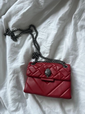 Röd quiltad handväska Kurt Geiger - Snygg röd quiltad handväska från Kurt Geiger med silverfärgad kedjerem och ikonisk örndetalj på locket. Väskan är i mjukt konstläder och har en kompakt, rektangulär form. Perfekt accessoar för att lyfta din outfit. Oanvänd. 