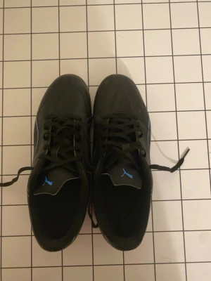 Svarta Puma sneakers med blå detaljer - Snygga svarta sneakers från Puma med blå logga på plösen och blå detaljer på sidan. Skorna har klassisk låg profil, snörning och ovandel i syntetmaterial. Perfekta för dig som vill ha en sportig och stilren look.