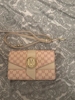 Beige rosa handväska  - Snygg beige handväska med guldigt M-emblem och justerbar axelrem. Väskan har ett mönster med M-loggor över hela ytan och detaljer i ljusbeige konstläder. Perfekt accessoar för att lyfta din outfit. För mer bilder/justering av pris skriv gärna❤️🤗