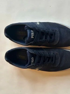 Marinblå sneakers från Polo Ralph Lauren - Snygga marinblå sneakers från Polo Ralph Lauren med klassisk låg profil. Skorna har vit sula, mörkblå ovandel i textil och ikoniska Polo-loggan broderad på sidan. Perfekta för en clean och stilren look.