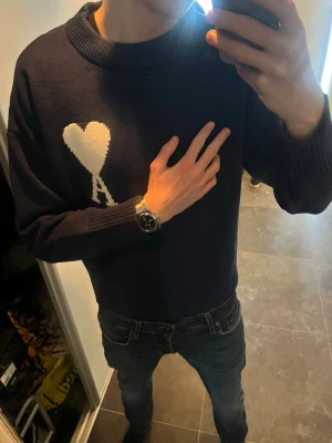 Ami Paris tröja  - Säljer en mörkblå stickad tröja från Ami Paris med deras ikoniska vita hjärta och A på bröstet. Tröjan har rund halsringning, ribbade muddar och en avslappnad passform. Perfekt för dig som gillar stilrena och trendiga plagg.