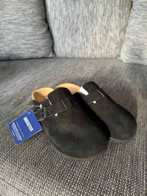 Svarta Birkenstock tofflor i mocka - Säljer ett par svarta Birkenstock tofflor i mjuk mocka med klassisk korksula och justerbart spänne. Modellen har öppen häl och ergonomisk fotbädd för extra komfort. Perfekta för dig som vill ha både stil och bekvämlighet.