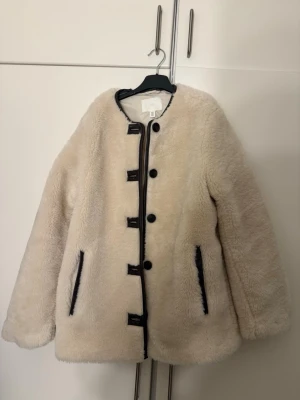 Beige teddyjacka från H&M XS - Supermjuk och fluffig beige teddyjacka från H&M i storlek XS. Jackan har svarta detaljer längs knäppningen och vid fickorna, rund halsringning och knappar framtill. Perfekt för att hålla sig varm och snygg under kyliga dagar.