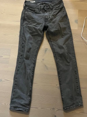 Grå Levi's jeans  - Säljer ett par grå jeans från Levi's med klassisk rak passform och fem fickor. Jeansen har bälteshällor, knappgylf och en liten röd Levi's-tagg på bakfickan. Materialet är denim i bomull och färgen är tvättad grå. Strl 28-30