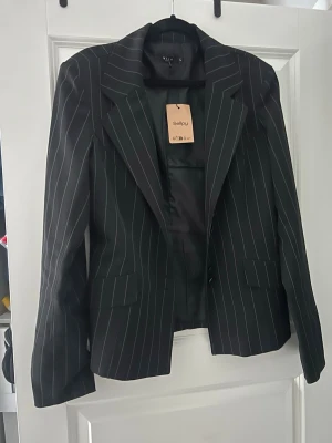 Svart kavaj med vita ränder från VILA - Snygg svart kavaj från VILA med tunna vita kritstrecksränder. Klassisk modell med breda slag, två framfickor med lock och enkelknäppning. Perfekt för en stilren och trendig look. Materialet är slätt och fodrat för extra komfort💕