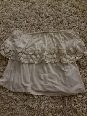 Vit offshoulder topp Gina Tricot - Supersöt vit offshoulder topp från Gina Tricot i storlek XS. Toppen har en bred volang med blommig spetsdetalj och är axelbandslös. Perfekt till sommaren och ger en romantisk vibe till din outfit.