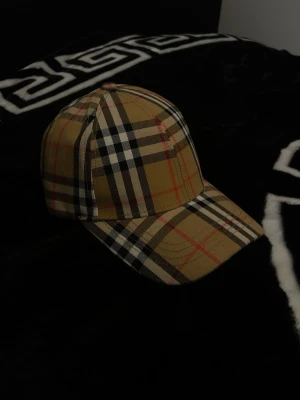 Burberry keps - Snygg keps från Burberry med det ikoniska rutmönstret i beige, svart, vitt och rött. Kepsen har böjd skärm och justerbar rem med metallspänne bak. Den är har endast prövats ett par gånger men är i helt nytt skick ställ gärna eventuella frågor eller prisförslag Mvh Clothes4you😊🤝