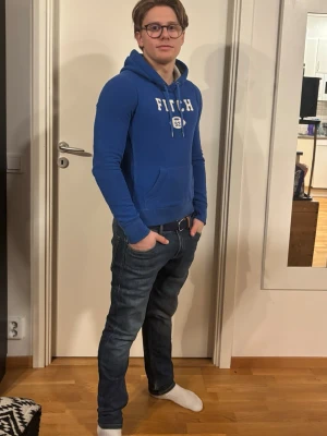 Blå hoodie från Fitch & acrombie - Snygg blå hoodie från Fitch med vit logga och tryck på bröstet. Klassisk känguruficka framtill och justerbar dragsko i huvan. Perfekt för en avslappnad och sportig stil. Mjuk bomullskänsla och ribbade muddar vid ärmslut och midja.
