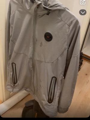 Ljusgrå vindjacka från Zanevs Canada - Ljusgrå vindjacka med huva från Zanevs Canada. Jackan har hel dragkedja framtill, två fickor med svarta dragkedjor och ett runt märke på bröstet. Tillverkad i ett lätt och vindtätt material, perfekt för blåsiga dagar.