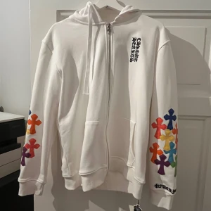 Vit hoodie Chrome Hearts  - Vit hoodie från Chrome Hearts med dragkedja, huva och stora fickor. Ärmarna är dekorerade med färgglada kors i blått, grönt, gult, orange, rött och lila. Snygg logga på bröstet och detaljer vid ärmslutet. Perfekt statement-plagg för dig som gillar streetwear.