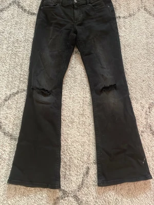 Svarta bootcut jeans med slitningar - Säljer ett par svarta bootcut jeans med slitningar över knäna och en mindre slitning på bakfickan. Jeansen har klassisk femficksmodell och knappgylf. Perfekta för dig som gillar en edgy och avslappnad stil.