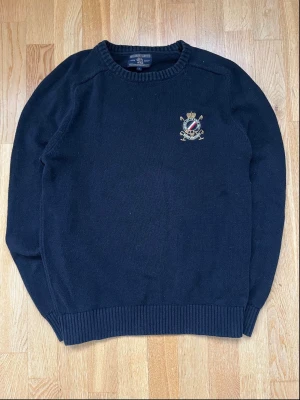 Mörkblå stickad tröja Morris Gents - Mörkblå stickad tröja från Morris Gents med rund hals och ribbade muddar. Tröjan har ett broderat emblem i guld, rött och blått på bröstet som ger en klassisk och preppy känsla. Perfekt för dig som gillar stilrena och tidlösa plagg.